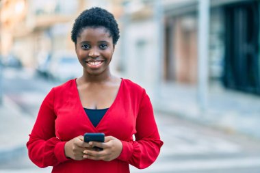 Genç Afrikalı Amerikalı kadın şehirde akıllı telefon kullanarak mutlu bir şekilde gülümsüyor..