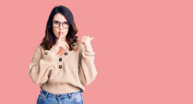 Güzel, genç, esmer, günlük kıyafetler ve gözlük takan bir kadın sessiz olmak istiyor. Parmakları dudaklarında, elleri yana doğru işaret ederek. sessizlik ve gizli konsept. 