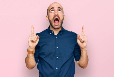 Gündelik giysiler giyen genç İspanyol adam şaşırmış ve şaşırmış bir şekilde yukarı bakıyor ve parmaklarıyla işaret ediyor ve kollarını kaldırıyor.. 