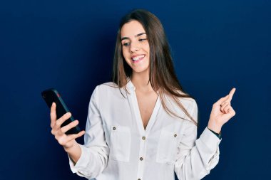 Genç esmer kız akıllı telefon kullanıyor. Masada oturmuş gülümsüyor. Mutlu bir şekilde işaret ediyor. 