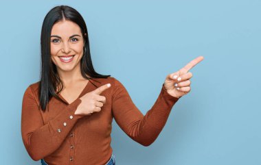 Gündelik kıyafetler giyen, gülümseyen ve kameraya bakan genç İspanyol kadın iki eliyle ve parmaklarıyla kenarı işaret ediyor.. 