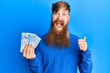 Genç İrlandalı kızıl saçlı adam Güney Afrika 'yı tutuyor. 100 rand' lık banknotlar başparmağıyla yan tarafı işaret ediyor. Ağzı açık gülümsüyor. 