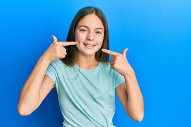 Güzel, esmer, günlük beyaz tişört giyen neşeli bir şekilde gülümseyen ve parmaklarıyla ve ağzıyla işaret eden küçük bir kız. diş sağlığı kavramı. 