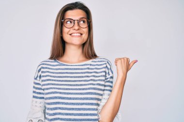 Güzel, beyaz bir kadın, günlük kıyafetler ve gözlük takıyor, gülümsüyor, gülümsüyor ve başparmağıyla yan tarafa bakıyor.. 