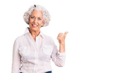 Yaşlı, gri saçlı, günlük kıyafetler giyen, gülümseyen, gülümseyen ve baş parmağıyla yüzünü gösteren bir kadın.. 