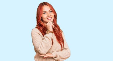 Genç kızıl saçlı kadın, sıradan bir kazak giyiyor kameraya kendinden emin bakıyor kollarını kavuşturmuş ve çenesini kaldırmış gülümsüyor. Olumlu düşünmek. 