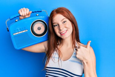 Genç kızıl saçlı kız elinde klasik radyoyla gülümsüyor mutlu ve pozitif, başparmağıyla mükemmel ve onay işareti yapıyor. 