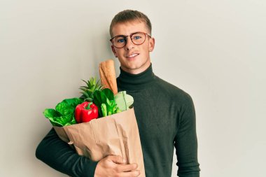 Elinde alışveriş poşeti olan beyaz bir genç. Pozitif ve mutlu görünüyor. Kendine güvenen bir gülümsemeyle dişlerini gösteriyor. 