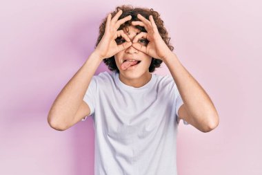 Yakışıklı, beyaz tişört giyen, dürbün gibi jest yapan, gözleri parmaklarından bakan genç bir adam. çılgın ifade. 