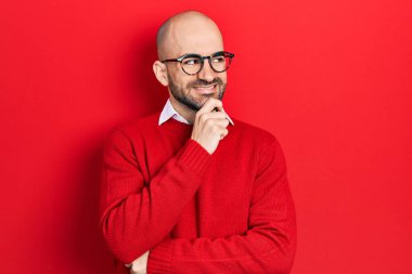 Genç kel bir adam günlük kıyafetler giyiyor ve elleri çenesinde gözlük takıyor soru sormayı düşünüyor, düşünceli bir ifade. Düşünceli bir yüzle gülümsüyor. şüphe kavramı. 