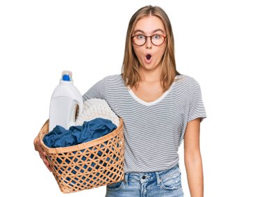 Çamaşır sepeti ve deterjan şişesi tutan güzel sarışın kadın şaşkınlık ve şaşkınlık içinde ağzı açık, inançsız bir yüz. 