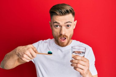 Elinde diş macunu ve bir bardak suyla diş fırçası tutan genç, kızıl saçlı bir adam şaşkınlık ve şaşkınlık dolu bir ifade, korku ve heyecanlı bir yüz ifadesiyle şoka girdi.. 