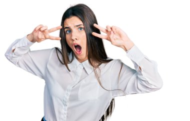 Genç esmer ergen barış jesti yapıyor. Gözleri kapalı. Şaşırmış ve şaşırmış bir ifade, korku ve heyecanlı bir yüz ifadesi var.. 