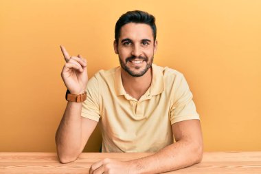 Masada oturan, yüzünde kocaman bir gülümseme olan, elleri ve parmaklarıyla kameraya bakan İspanyol asıllı genç adam.. 