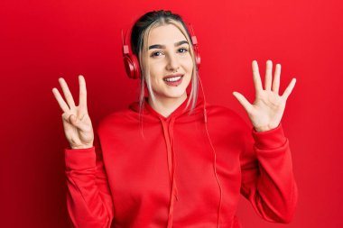 Genç modern kız spor kıyafetleri giyiyor ve kulaklık takıyor. Sekiz numaralı parmaklarıyla işaret ediyor. Kendine güvenen ve mutlu bir şekilde gülümsüyor.. 