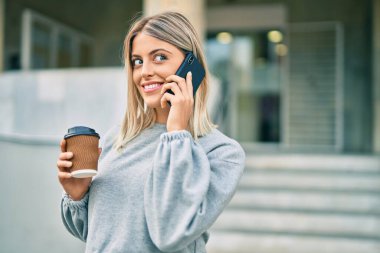 Genç sarışın kız akıllı telefondan konuşuyor ve şehirde kahve içiyor.