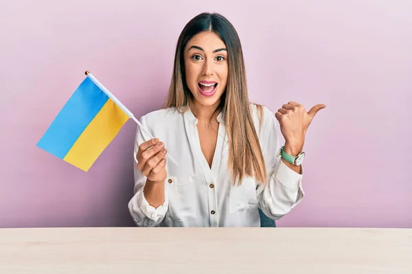 若いですヒスパニック女性保持Ukraine Flag座っています上のテーブル指す親指へザ側笑顔幸せとともにオープン口 — ストック写真
