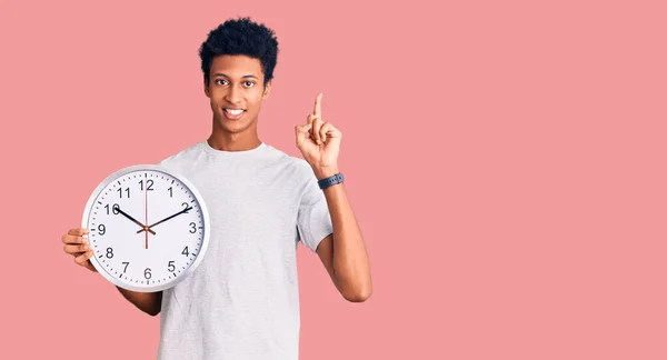 若いアフリカ系アメリカ人の男を持っている大きな時計に驚いてアイデアや質問を指差す指で幸せな顔 ナンバーワン — ストック写真