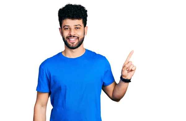 年轻的阿拉伯男子 留着胡子 穿着休闲的蓝色T恤 脸上挂着大大的笑容 手指头指向侧面看相机 — 图库照片