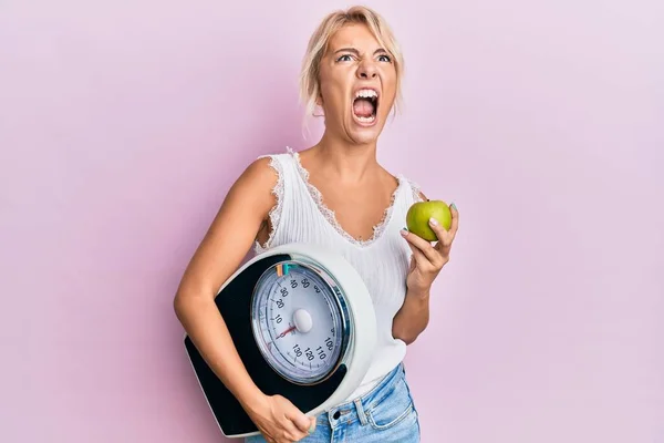 Young Blonde Girl Holding Weight Machine Green Apple Angry Mad — Zdjęcie stockowe