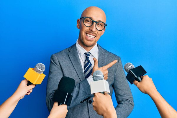 Bald man with beard being interviewed by reporters holding microphones smiling cheerful pointing with hand and finger up to the side 