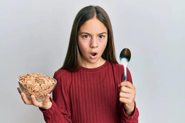 美しいブルネットの女の子食べる健康な全粒粉穀物保持スプーンで衝撃的な顔 懐疑的で皮肉 驚きでオープン口 — ストック写真