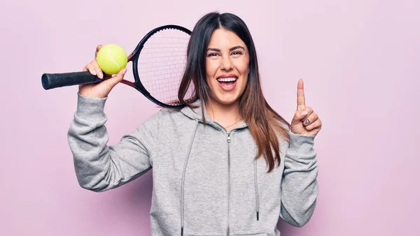 若い美しいスポーツ女性を使用してテニスをプレイラケットとボールピンクの背景笑顔でアイデアや質問を指差す指で幸せな顔 ナンバーワン — ストック写真