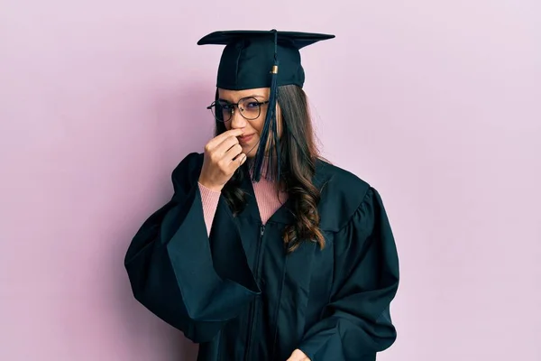 卒業キャップと式のローブを着た若いヒスパニック系の女性は 臭いと嫌な 耐えられない匂いを嗅ぎ 鼻の上に指で息を保持します 臭いが悪い — ストック写真
