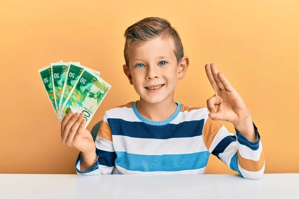 Criança Caucasiana Adorável Segurando Israel Shekels Sentado Mesa Fazendo Sinal — Fotografia de Stock