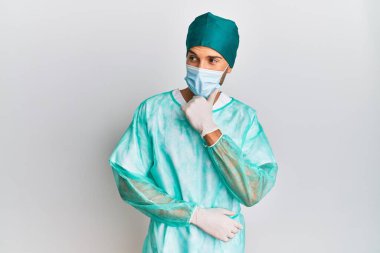 Cerrah üniforması giyen ve elleri çenesinde tıbbi maske takan soru soran genç yakışıklı bir adam, düşünceli bir ifade. Düşünceli bir yüzle gülümsüyor. şüphe kavramı. 