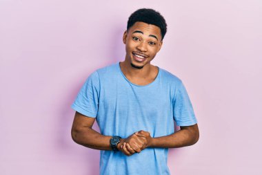 Genç Afro-Amerikalı adam, günlük mavi tişörtler giyip ellerini kavuşturmuş ve mutlu bir şekilde gülümsüyor. Başarı ve iyimserlik 