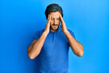 Latin asıllı genç bir adam, günlük mavi tişört giyiyor, elleri kafasında, stresten baş ağrısı çekiyor. migren ağrısı. 