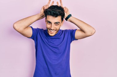 Gündelik tişört giyen genç İspanyol adam komik ve çılgın poz veriyor parmakları kafasında tavşan kulağı, neşeli bir gülümseme 