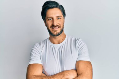 Gündelik beyaz tişört giyen genç İspanyol adam kameraya bakarken kollarını kavuşturup gülümsüyor. pozitif kişi. 