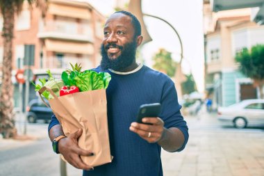 Sakallı Afrikalı Amerikalı adam elinde süpermarketten alınmış kağıt torbalarla akıllı telefon kullanıyor.