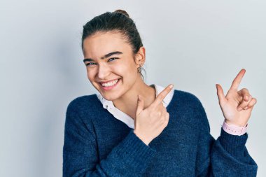 Gündelik giysiler giymiş genç beyaz bir kız gülümseyip kameraya bakıyor ve iki eliyle ve parmaklarıyla kenarı işaret ediyor.. 