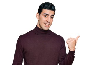 Yakışıklı, balıkçı yaka kazak giyen, gülen yüzlü gülümseyen ve baş parmağıyla yüzünü gösteren bir İspanyol.. 