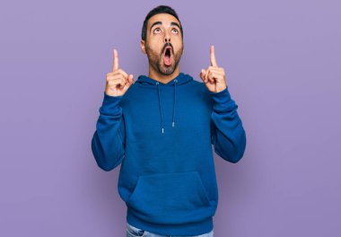 Gündelik giysiler giyen genç İspanyol adam şaşırmış ve şaşırmış bir şekilde yukarı bakıyor ve parmaklarıyla işaret ediyor ve kollarını kaldırıyor.. 