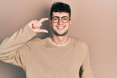 Günlük kıyafetler giyen, gözlüklü, gülümseyen, kendine güvenen, el hareketi yapan, parmaklarıyla işaret yapan ve kameraya bakan genç İspanyol bir adam. ölçüm kavramı. 