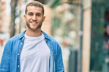 Genç beyaz adam gülümseyerek şehirde mutlu mesut duruyor..