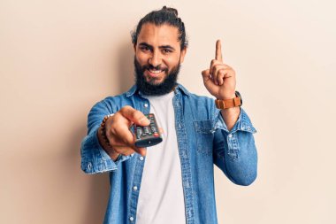 Genç Arap adam elinde uzaktan kumandayla bir fikir ya da soruyla gülümsüyor ve mutlu bir yüzle işaret ediyor. 