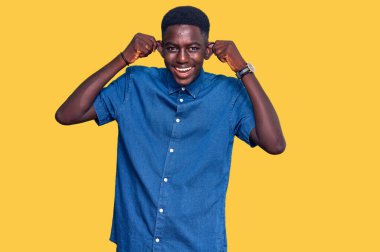 Genç Afro-Amerikan bir adam günlük kıyafetler giyiyor gülümsüyor kulakları parmaklarıyla çekiyor, komik bir jest. deneme sorunu 