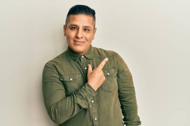 Gündelik kıyafetler giyen genç latin adam, el parmağı ile yan tarafı işaret ederek ciddi ve sakin bir yüz ifadesi sergiliyor. 