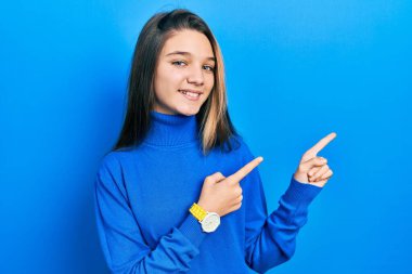 Balıkçı yaka kazak giyen, gülümseyen ve kameraya bakan genç esmer kız iki eliyle ve parmaklarıyla yan tarafı işaret ediyor.. 