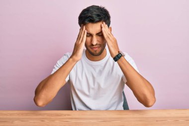 Masada oturan sıradan beyaz t-shirt giyen genç yakışıklı adam acı ve migrenden dolayı çaresiz ve stresli bir şekilde baş ağrısı çekiyor. Eller başının üstünde.. 
