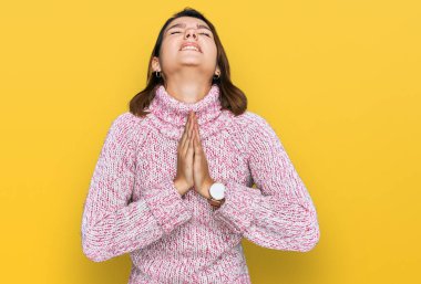 Kışın yün kazak giyen genç beyaz kız elleri bağlı yalvarıyor ve dua ediyor. Yüzünde umut ifadesi var. Çok duygusal ve endişeli. Yalvarıyor. 