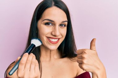 Makyaj fırçası tutan, yüzü kızaran, mutlu ve pozitif gülümseyen, başparmağı mükemmel ve onay işareti yapan genç İspanyol kadın. 