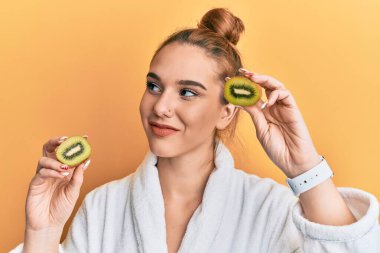 Bornoz giyen genç sarışın kadın elinde taze kivi, gülümsüyor ve yan tarafa bakıyor.. 