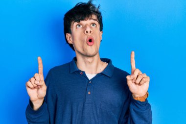 Yakışıklı hippi genç adam mavi arka planda mahsur kalmış şaşkın şaşkın yukarı bakıyor ve parmaklarıyla işaret ediyor ve kollarını kaldırıyor.. 