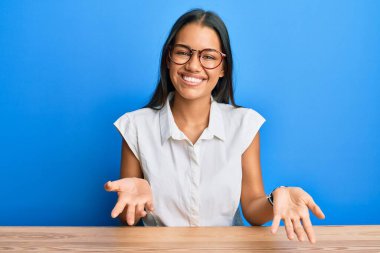 Güzel İspanyol kadın gündelik giysiler içinde masada oturmuş neşeli bir şekilde kollarını açıp gülümsüyor. Hoş geldin, olumlu ve kendinden emin selamlar. 
